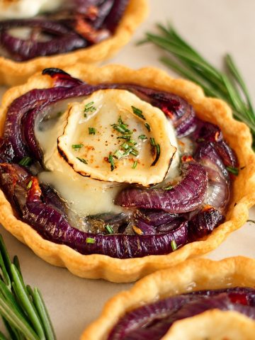 Gros plan sur une tartelette aux oignons rouges garnie de chèvre fondu et de romarin.