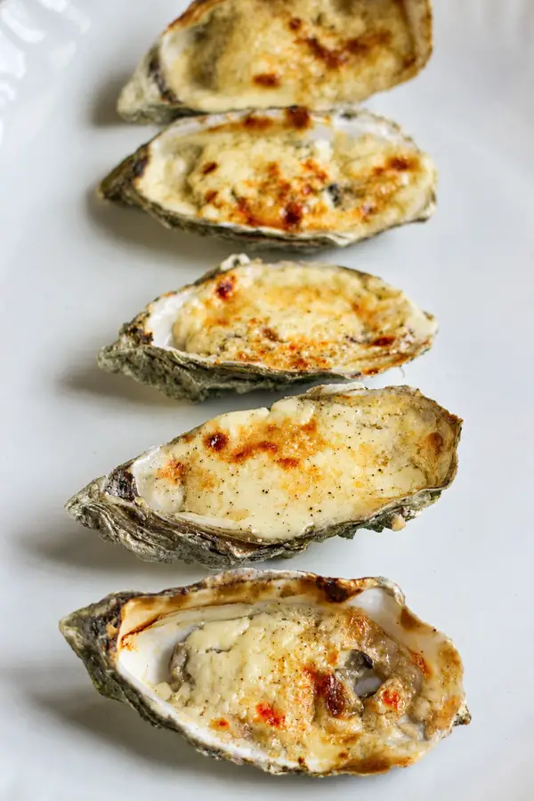 Cinq huîtres gratinées au four, alignées sur une assiette blanche avec une garniture crémeuse et dorée.
