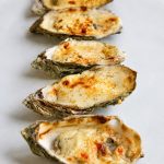 Gros plan sur cinq huîtres gratinées au four, recouvertes d’une garniture crémeuse au parmesan joliment dorée.