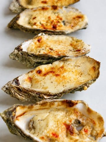 Gros plan sur cinq huîtres gratinées au four, recouvertes d’une garniture crémeuse au parmesan joliment dorée.