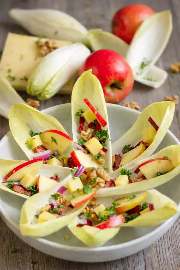 Salade d'endives en forme de fleur sur assiette blanche aux pommes, oignons rouges, comté, noix et lardons.