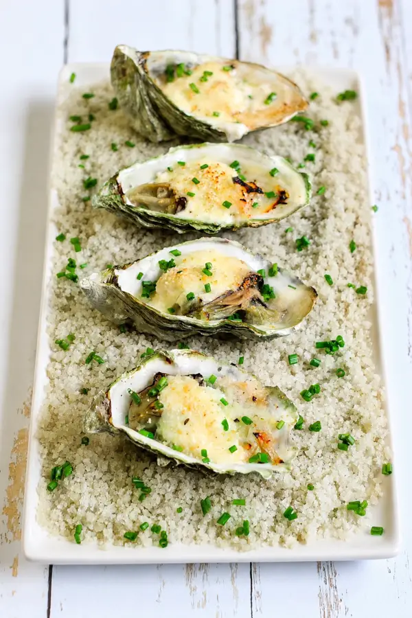 Quatre huîtres gratinées au champagne, garnies de chapelure dorée et de ciboulette, présentées sur un lit de gros sel.